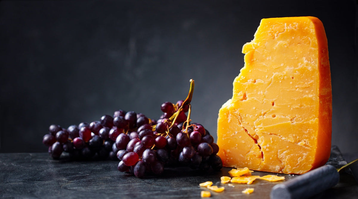 A wedge of orange cheddar cheese beside a bunch of dark red grapes on a slate surface.