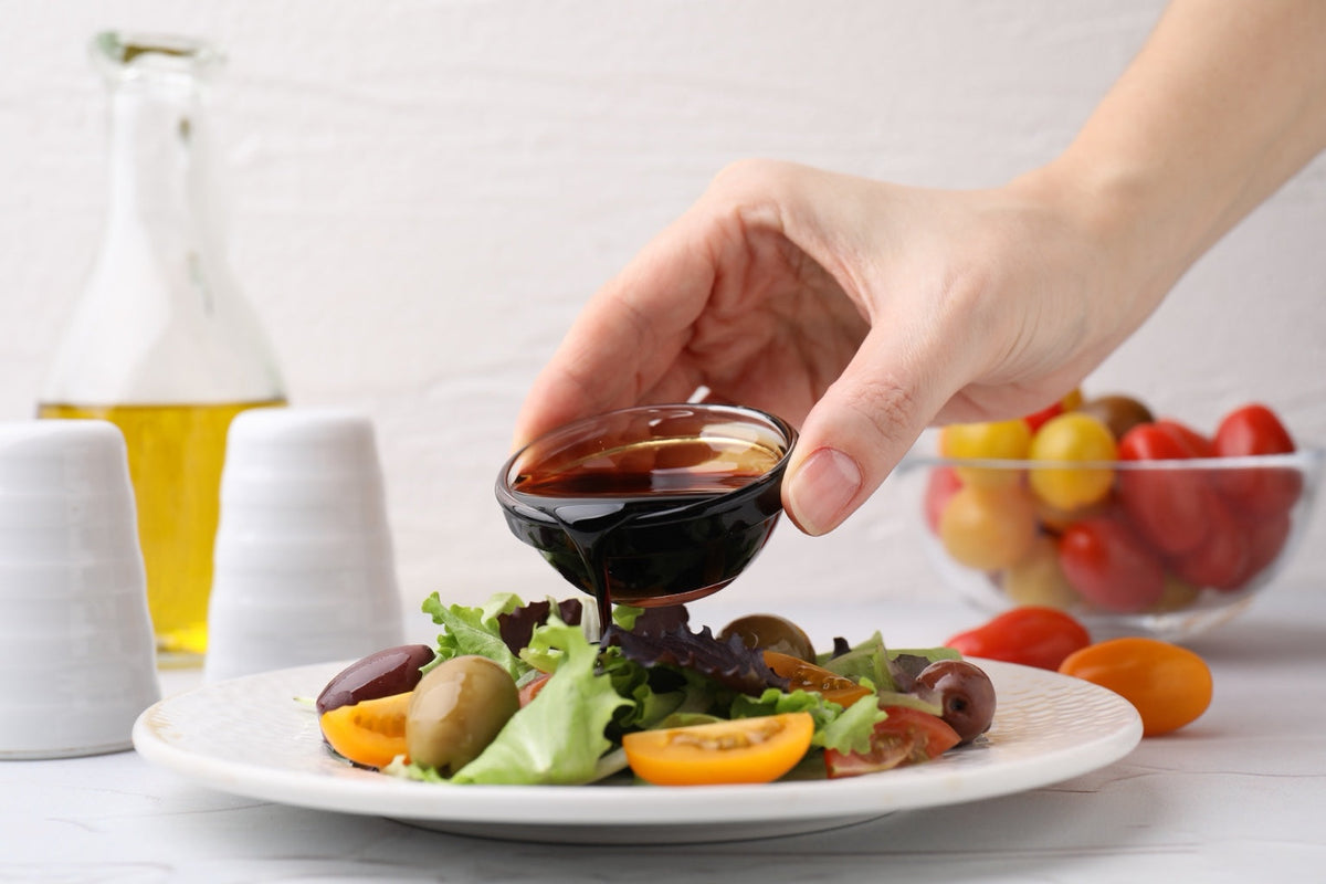A hand pours rich Italian balsamic vinegar over a fresh salad with tomatoes and olives on white porcelain
