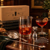 A Bar Above stainless steel stirred cocktail set and gift box on a dark wood bar; serving glass and drinks are styling props.