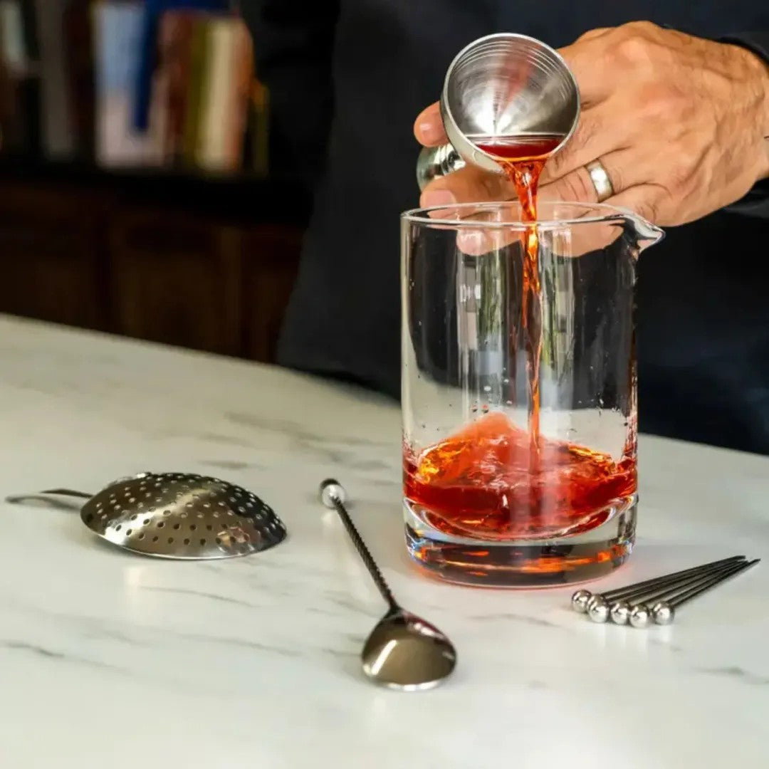 Bell jigger pouring into the A Bar Above mixing glass, with stainless steel julep strainer, spiral bar spoon and cocktail picks.