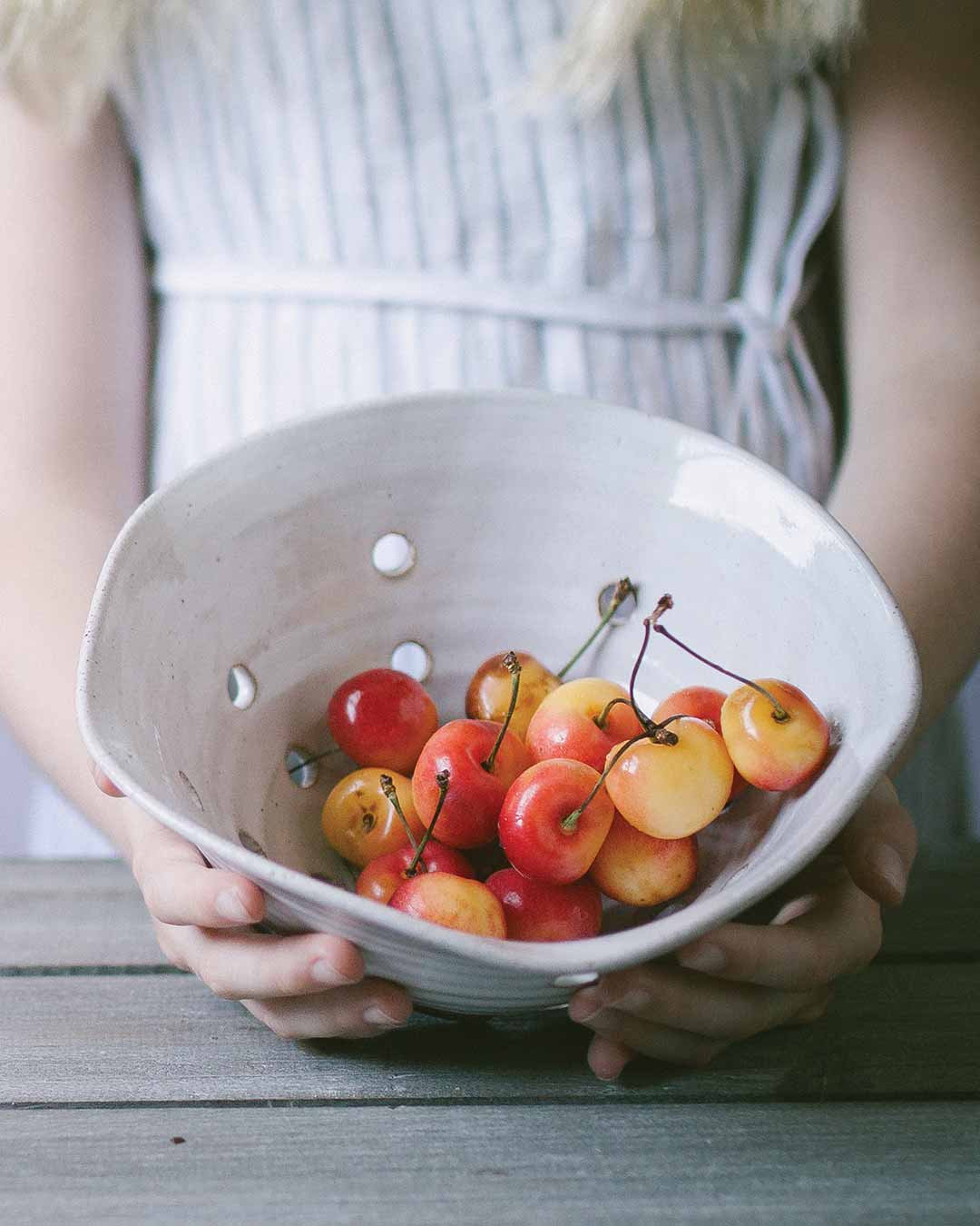 White Farmhouse Pottery Windrow Berry Bowl filled with cherries, held to show the hand-carved drainage holes; cherries not included.