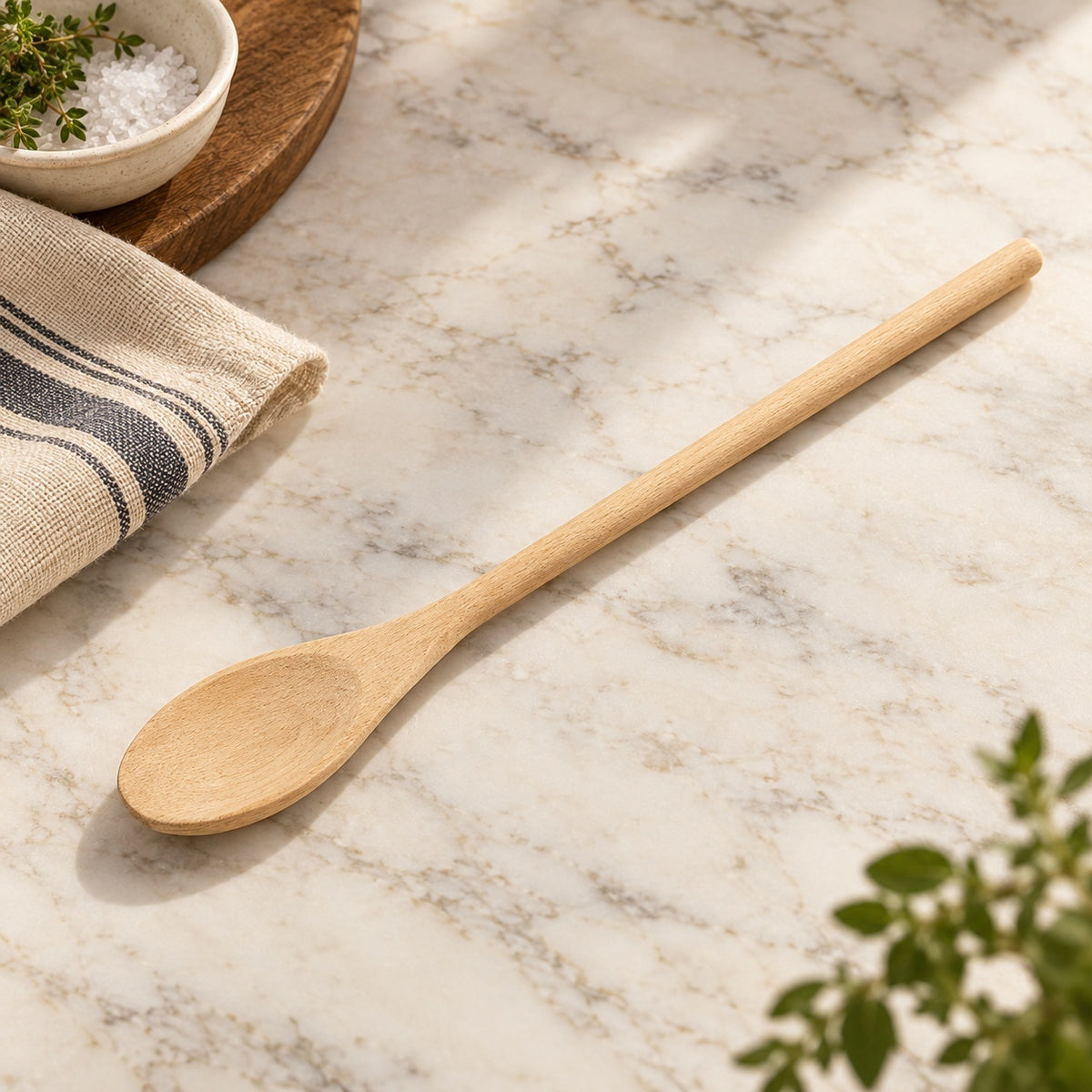 Fox Run 12-inch oval beechwood spoon on pale marble, styled with striped linen, herbs and a ceramic salt bowl.